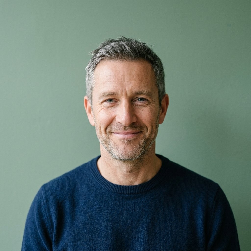 Portrait of Callum Ashworth, a man in his late forties in a navy jumper, smiling in front of a plain sage green wall